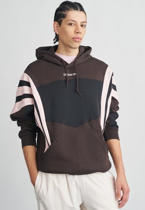 Joven con cabello trenzado que lleva una sudadera con capucha Adidas marrón y negra con rayas rosas y pantalones blancos, de pie con las manos en los bolsillos.