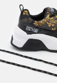 Baskets épaisses noires et blanches avec motif baroque doré, logo "Versace Jeans Couture" sur le talon et la semelle, et lacets noirs avec détails blancs.