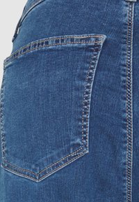 Primer plano de unos jeans de mezclilla azul que destacan un bolsillo trasero con un diseño bordado y hilo en contraste. La textura muestra un acabado suave.