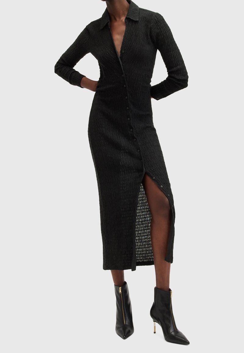 Femme portant une longue robe noire texturée à boutons avec une fente haute, associée à des bottines noires à talons hauts avec des fermetures éclair dorées.