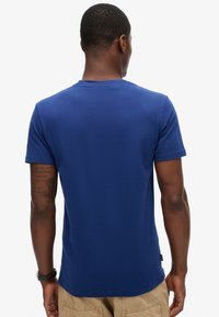 Camiseta de manga corta azul marino hecha de algodón, con cuello redondo y una textura suave. Muestra un tatuaje en el brazo derecho.