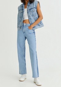 Veste en denim bleu clair sans manches, associée à un jean taille haute bleu clair. Des baskets blanches créent un contraste avec la tenue.