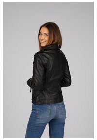 Schwarze Lederjacke mit tailliertem Design, Schulterepauletten und Reißverschlussakzenten. Modell trägt blaue Jeans und blickt mit dem Rücken zum Betrachter.