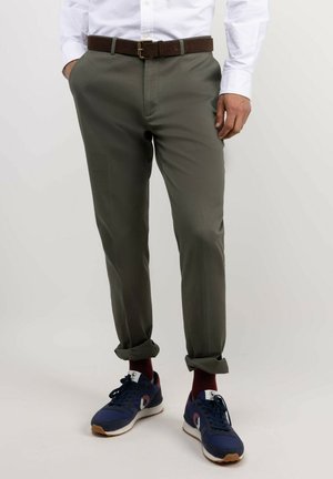 Hombre con pantalones verdes oliva remangados, cinturón marrón, camisa blanca, calcetines rojos y zapatillas azul marino, de pie con las manos en los bolsillos.