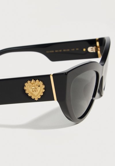 Μαύρα γυαλιά ηλίου cat-eye με σκουρόχρωμους φακούς και χρυσό σήμα σε σχήμα καρδιάς στον βραχίονα, με περίτεχνη διακόσμηση και μικρό λογότυπο.