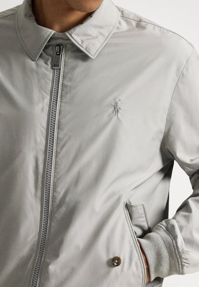 Ανοιχτό γκρι μπουφάν με φερμουάρ μπροστά, μυτερό γιακά και μανσέτες με ριμπ ύφανση. Διαθέτει διακριτικό κεντητό λογότυπο και τσέπη στο πλάι με κουμπί.