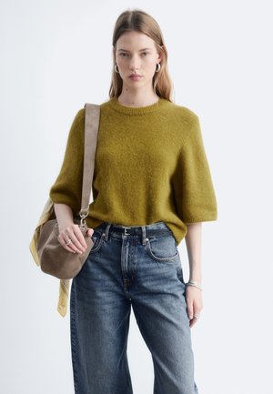 Femme portant un pull duveteux vert olive, un jean bleu taille haute, des boucles d'oreilles créoles argentées, et portant un sac à bandoulière en daim beige avec une écharpe jaune.