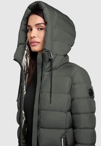 Pufferjack in olijfgroen met een grote capuchon, gequiltde textuur, rits aan de voorkant en zwarte hardware accenten. Van binnen gevoerd met zilver materiaal.