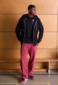Sudadera negra con cremallera y cordones blancos, camiseta negra, pantalones rojos holgados y zapatillas negras/rojas. Tela suave, corte relajado, detalles mínimos.