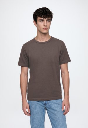 Jeune homme aux cheveux bouclés foncés, portant un t-shirt marron uni à manches courtes et un jean bleu clair, regardant légèrement vers la droite.