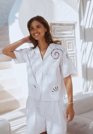 Femme souriante en chemise blanche à manches courtes et short avec des motifs découpés en spirale et en éventail, debout près d'escaliers ensoleillés et de murs blancs.