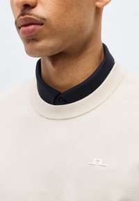 Κοντινό πλάνο ενός άνδρα που φοράει μπεζ πουλόβερ με στρογγυλή λαιμόκοψη πάνω από ένα μαύρο πουκάμισο με γιακά, με ένα μικρό λευκό λογότυπο στο στήθος του πουλόβερ.