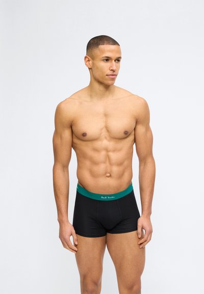 Μυώδης άνδρας που φοράει μαύρα μπόξερ με πράσινη ζώνη μέσης και την επιγραφή «Paul Smith», στέκεται και κοιτά προς τα δεξιά, σε απλό λευκό φόντο.
