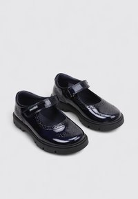 Un par de zapatos Mary Jane negros y brillantes con detalles perforados, suelas de goma gruesas y correas ajustables de Velcro sobre un fondo gris claro.