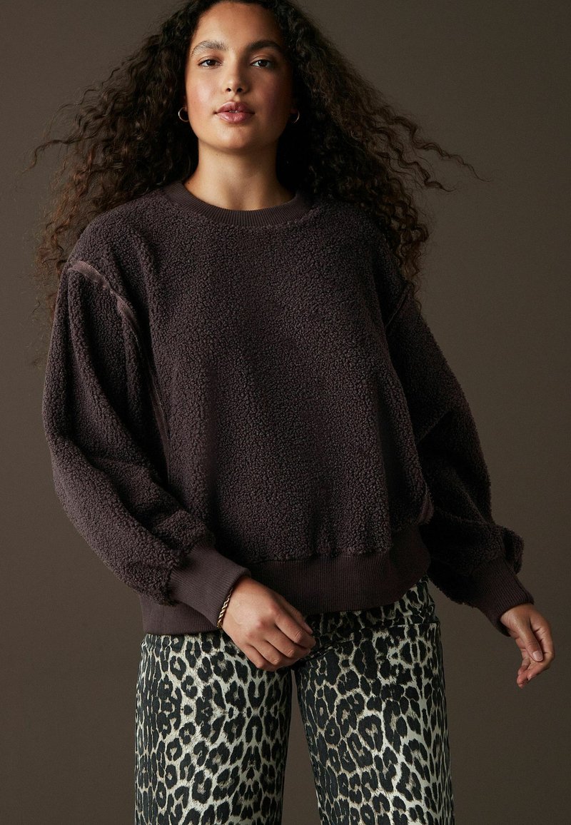 Brun fleece-tröja med texturerad yta, ribbade ärmar och nederkant; ihop med byxor i krämfärg och svart leopardmönster.