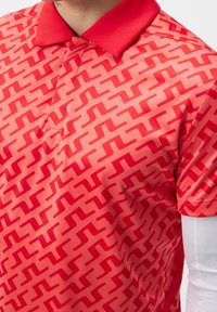 Camisa polo roja con un patrón geométrico de zigzag, hecha de tela suave. Presenta un cuello clásico y mangas cortas, acentuadas por colores contrastantes.