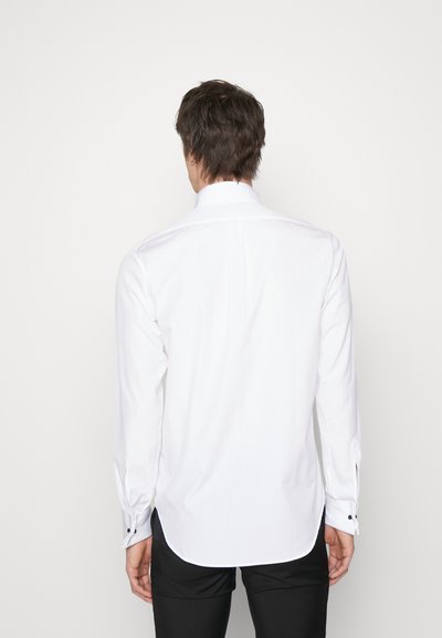 Chemise blanche à manches longues avec un col rabattu, ourlet arrondi, poignets à boutons et une texture lisse, accompagnée de pantalons noirs.