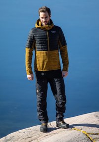 Black and mustard insulated jacket with a hood, textured body, and smooth sleeves, paired with black pants and sturdy black boots.