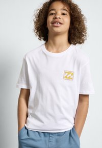 T-shirt en coton blanc à manches courtes, avec un petit logo jaune et bleu dans un design carré sur la zone du côté gauche de la poitrine.