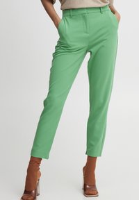 Un modello posa in pantaloni verdi vivaci, abbinati a scarpe con tacco alte marroni e calze rigate marroni coordinate, su uno sfondo bianco.