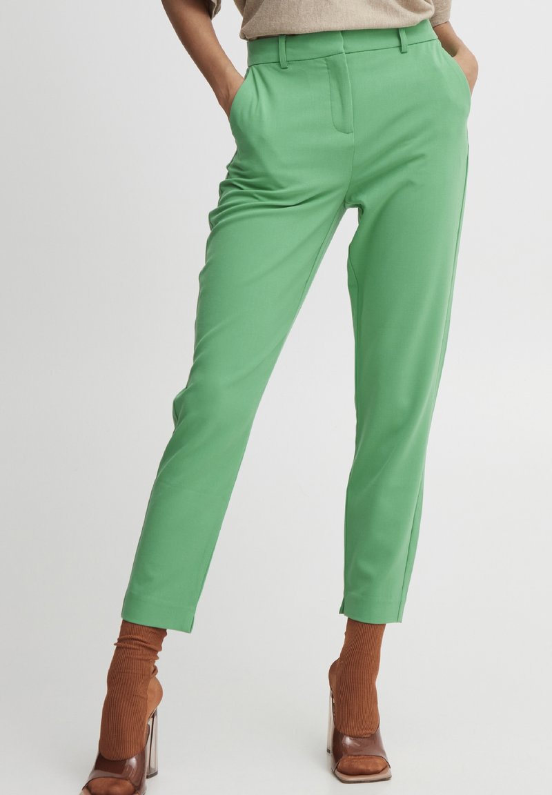 Un modello posa in pantaloni verdi vivaci, abbinati a scarpe con tacco alte marroni e calze rigate marroni coordinate, su uno sfondo bianco.