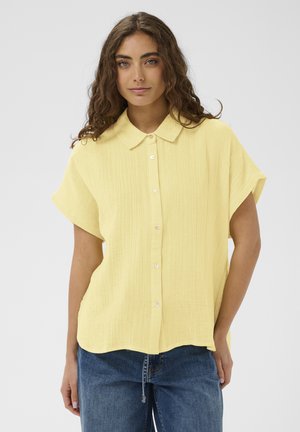 Žena s dlhými kučeravými vlasmi, oblečená v svetložltej krátkosleevej košeli na zapínanie a modrých džínsoch, stojaca pred jednoduchým pozadím.