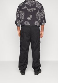 Henkilö, joka on pukeutunut mustiin cargo-housuihin ja mustaan puseroon, jossa on valkoisia paisley-kuvioita, seisoo yksinkertaista valkoista taustaa vasten.