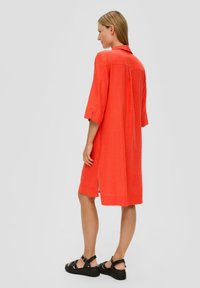 Rotes Leinenkleid mit lockerem Schnitt, aufgekrempelten Ärmeln, seitlichen Schlitzen und einem Kragen. Getragen mit schwarzen Riemchensandalen vor einem neutralen Hintergrund.