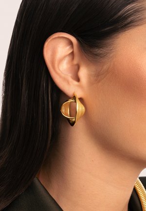 Boucles d'oreilles dorées présentant un design en spirale avec des textures superposées et une forme ovale ouverte, accrochées à un teint de peau neutre.