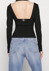 Top noir à manches longues avec un décolleté profond dans le dos, associé à un jean en denim clair avec deux poches arrière et une coupe légèrement ample.