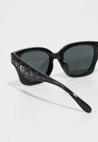 Lunettes de soleil noires en forme de chat avec des verres gris, finition brillante, branches noires texturées avec un accent doré.