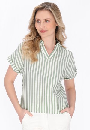 Kvinde med bølget blondt hår iført en kortærmet grøn og hvid stribet skjorte med lodrette striber og hvide bukser, hænderne i lommerne, kigger mod højre.