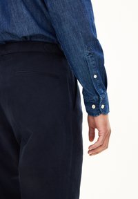 Donkerblauwe denim overhemd met lange mouwen en knoopmanchetten, gecombineerd met marineblauwe stoffen broek. Opvallende gladde textuur en slim-fit.