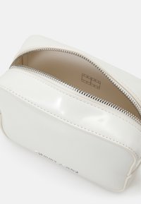 Bílá kosmetická taška z patentované kůže se zipovým uzávěrem, obdélníkový tvar a embosované logo. Hladký povrch a stříbrný zip.