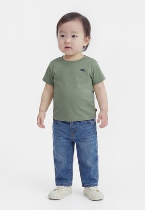 Peuter staat, draagt een groen Levi's T-shirt, blauwe spijkerbroek en witte sneakers tegen een effen witte achtergrond.