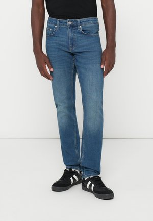 Blue jeans with a straight-leg cut, light fading, five-pocket design, metal hardware, and paired with black sneakers featuring white stripes.