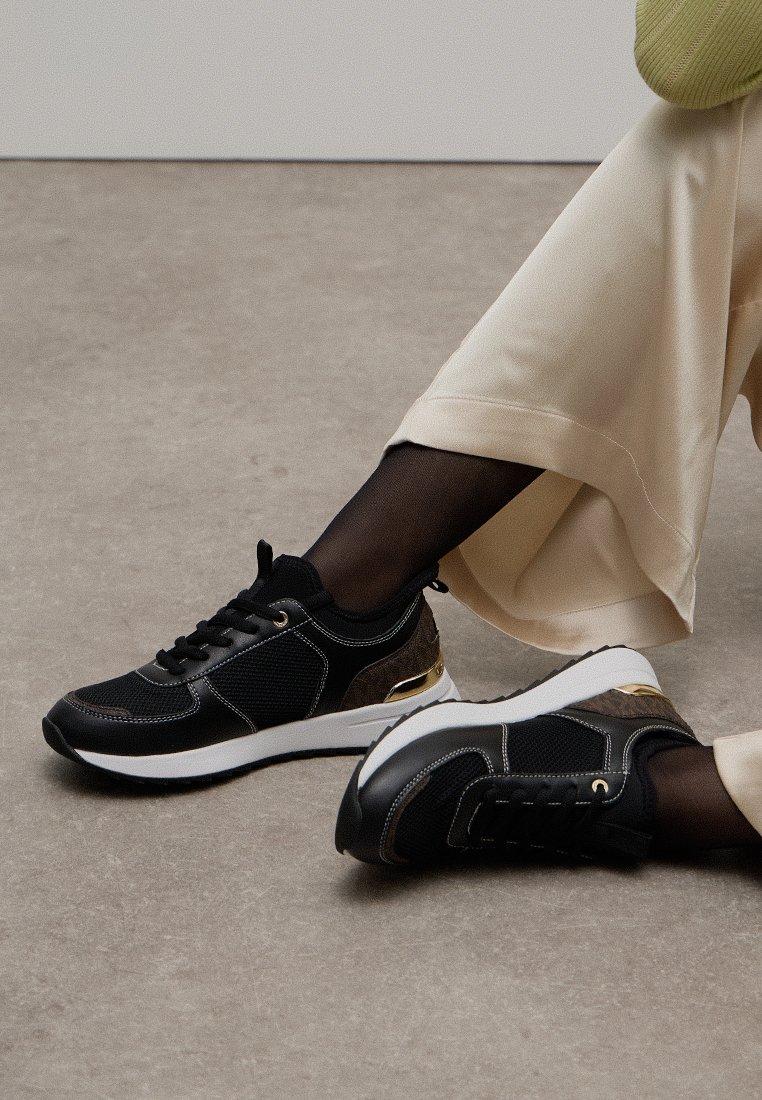 Persona vestida con pantalones beiges y zapatillas negras con suelas blancas, sentada en un suelo gris con una pierna cruzada sobre la otra.