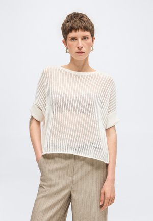 Lühikeste pruunide juustega naine, kes kannab kuldseid rõngaskõrvarõngaid, lahtist valget kootud topi rinnahoidja kohal ning beežikaid triibulisi pükse, käed taskutes.