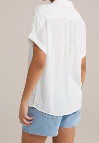 White short-sleeve shirt with a textured fabric, button-down collar, and a curved hem, paired with light blue denim shorts.