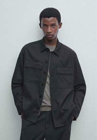 Giacca nera a bottoni realizzata in tessuto a trama, con due tasche sul petto e un design con colletto. Indossata sopra una camicia grigia con una texture liscia.