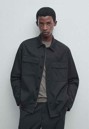 Black button-up jacket made of woven fabric, featuring two chest pockets and a collared design. Worn over a gray shirt with a smooth texture.