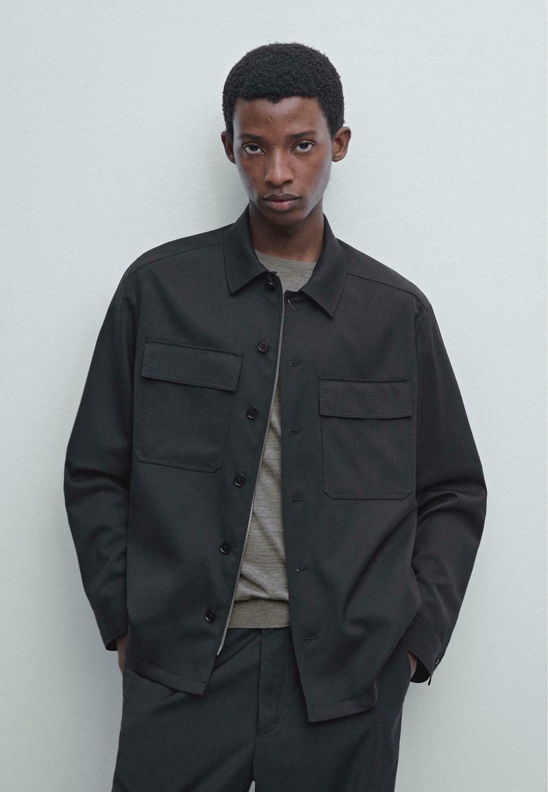 Giacca nera a bottoni realizzata in tessuto a trama, con due tasche sul petto e un design con colletto. Indossata sopra una camicia grigia con una texture liscia.