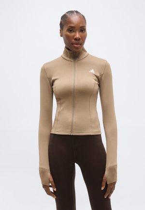 Kvinde iført en tætsiddende beige lynlåstrøje med hule til tommelfingrene og mørkebrune leggings, stående mod en ensfarvet lys baggrund.