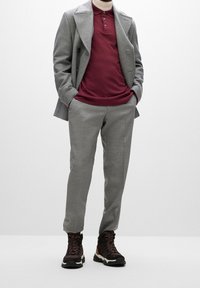 Grau wollener Übermantel, bordeauxrotes Polohemd, hellgraue maßgeschneiderte Hose und braune Wanderschuhe mit weißen Sohlen und bordeauxfarbenen Schnürsenkeln.
