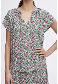 Haut fleuri en tissu léger, avec un col en V, des manches courtes, et un fond bleu pastel orné de fleurs rouges, roses et vertes.