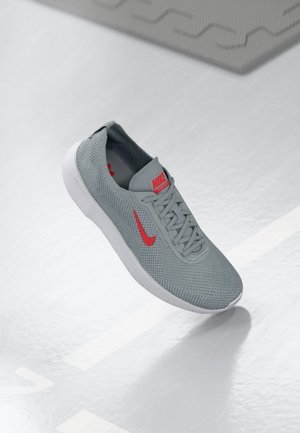 Chaussure de course Nike grise avec logo rouge et semelle blanche flottant au-dessus d'une surface blanche texturée.