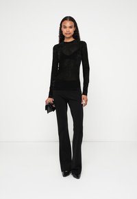 Naine seisab valge tausta ees, kandes musta siidist pikkade varrukatega toppi, musti pükse, teravapea kingi ning hoides käes väikest musta klambrit.