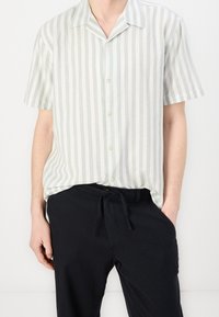 Short-sleeve, green-striped shirt in lightweight fabric, featuring a button-down front and a relaxed fit, paired with black drawstring pants.