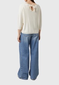 Kremowa dzianinowa bluzka z wycięciem w kształcie litery V z tyłu i ozdobną kokardą, zestawiona z jasnoniebieskimi szerokimi dżinsami. Gładka tekstura na całej powierzchni.