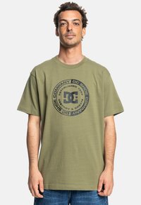 Olivegrüne Baumwoll-T-Shirt mit schwarzem runden Logo und dem Text "DC Shoe Company" sowie dem Gründungsjahr. Kurze Ärmel und entspannter Schnitt.
